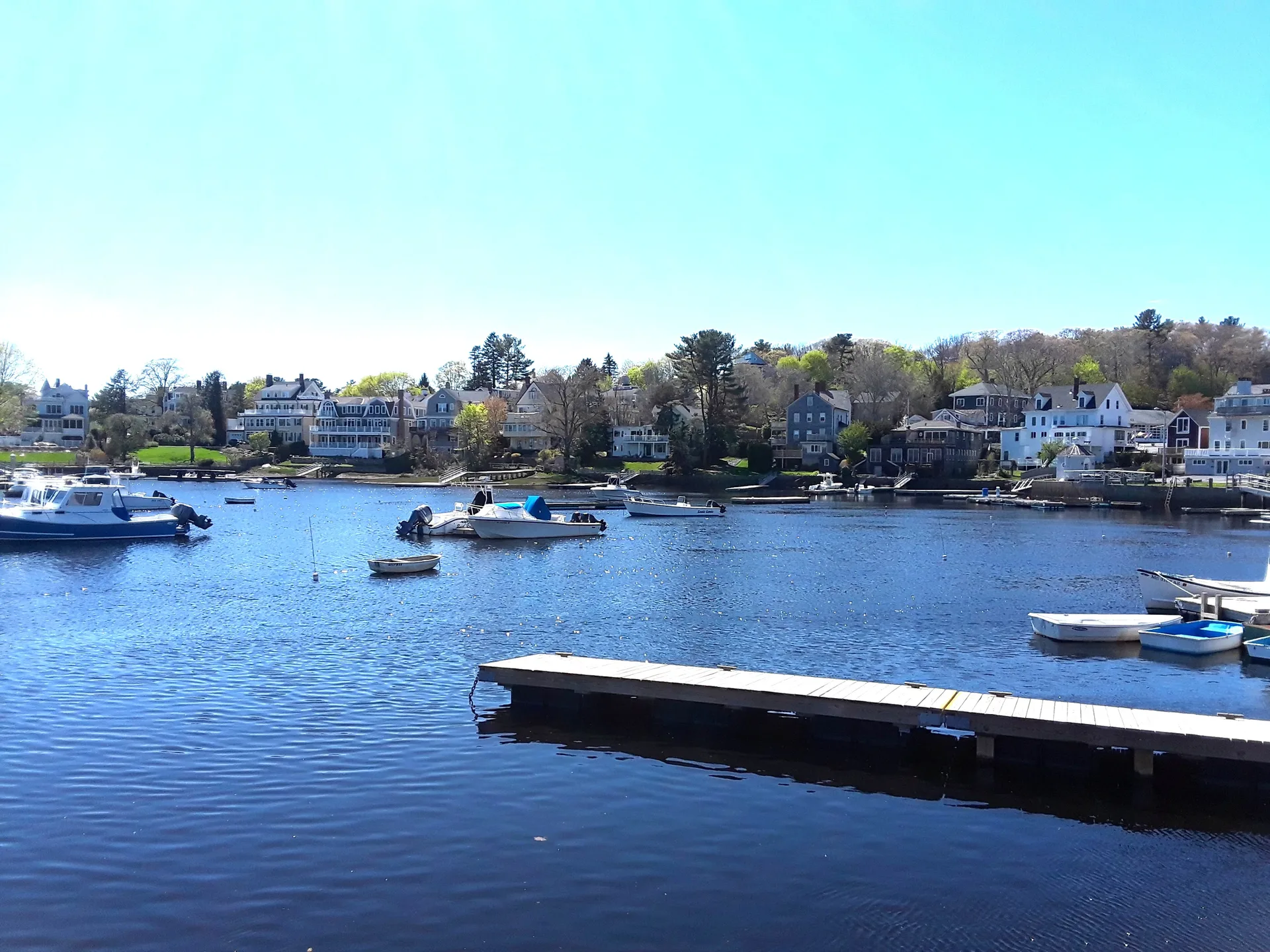 Manchester-by-the-Sea harbor waterfront in clear May daylight, boats moored along wooden docks with homes on the shore behind