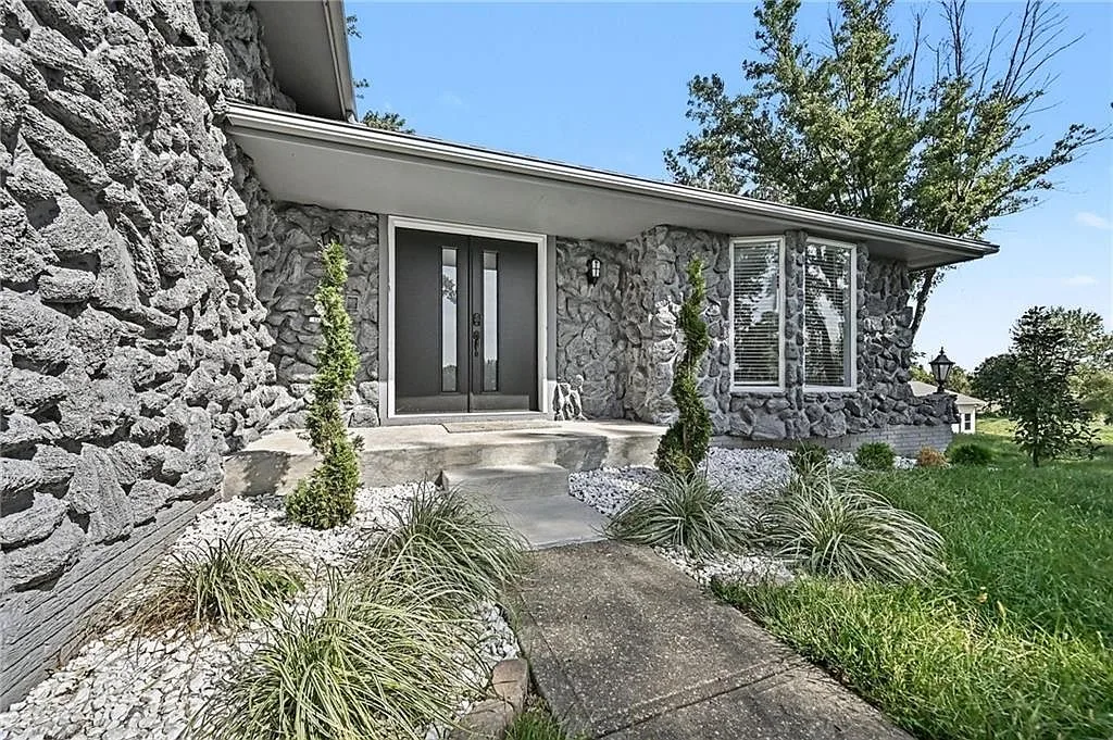 Ground level view of the stone front facade, dark double doors and landscaped entry with evergreens