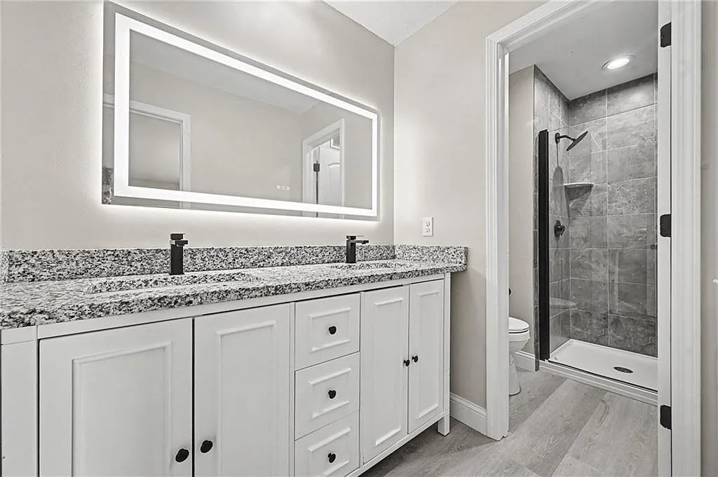 Renovated bath with dual white vanities, granite counter, matte black faucets and backlit mirror