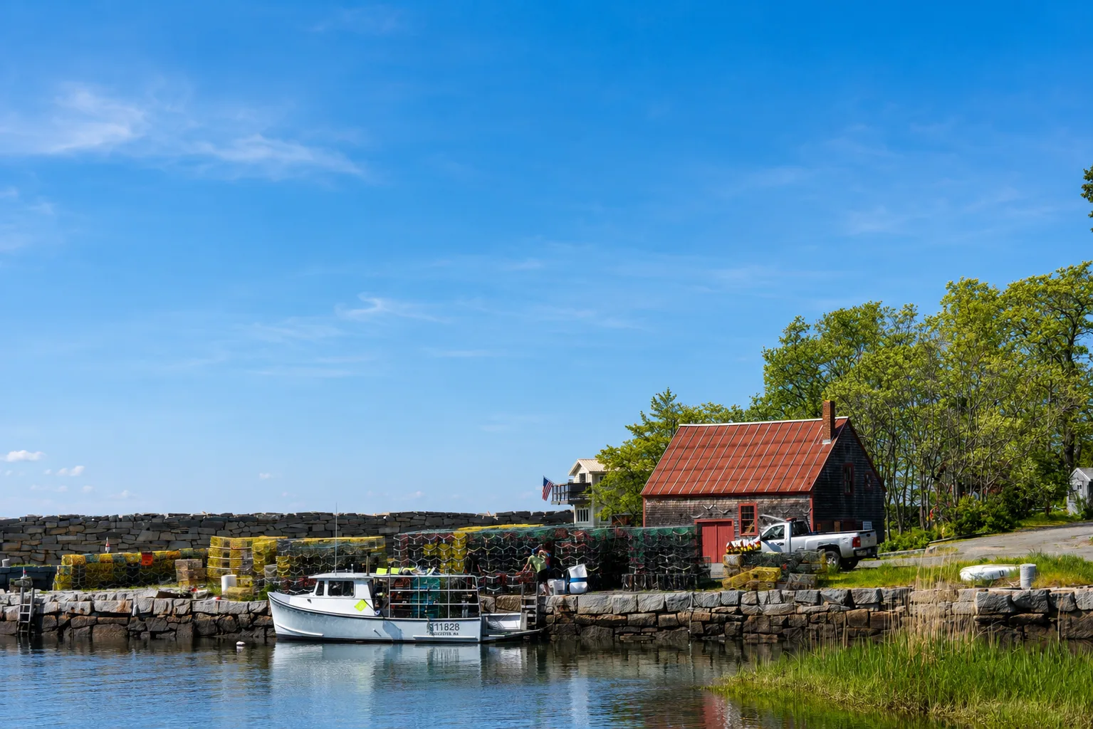 A weathered shingle building with a red roof beside a fishing boat in a Rockport-style harbor