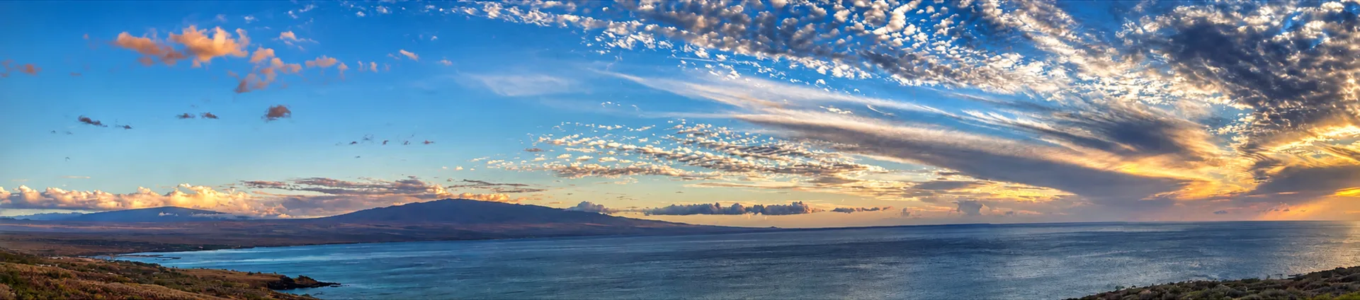 A wide panoramic sunset over the Kohala Coast of Hawaiʻi Island, with the coastline, mountains, and ocean spanning the horizon
