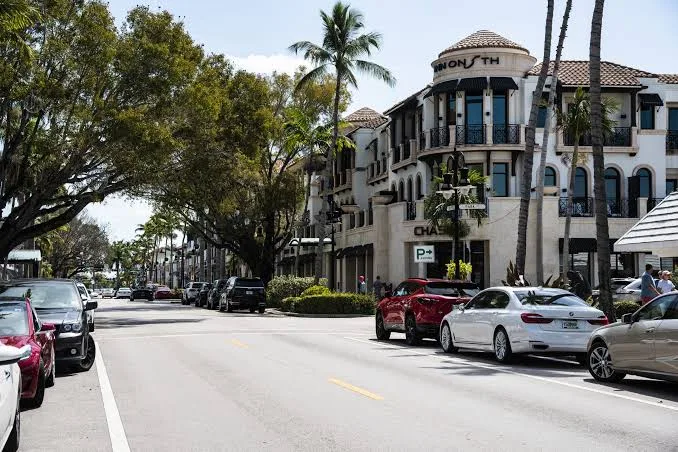 Daytime view of a Mediterranean-style commercial street in Olde Naples with stucco storefronts, a clay tile roof, and palm trees along the sidewalk