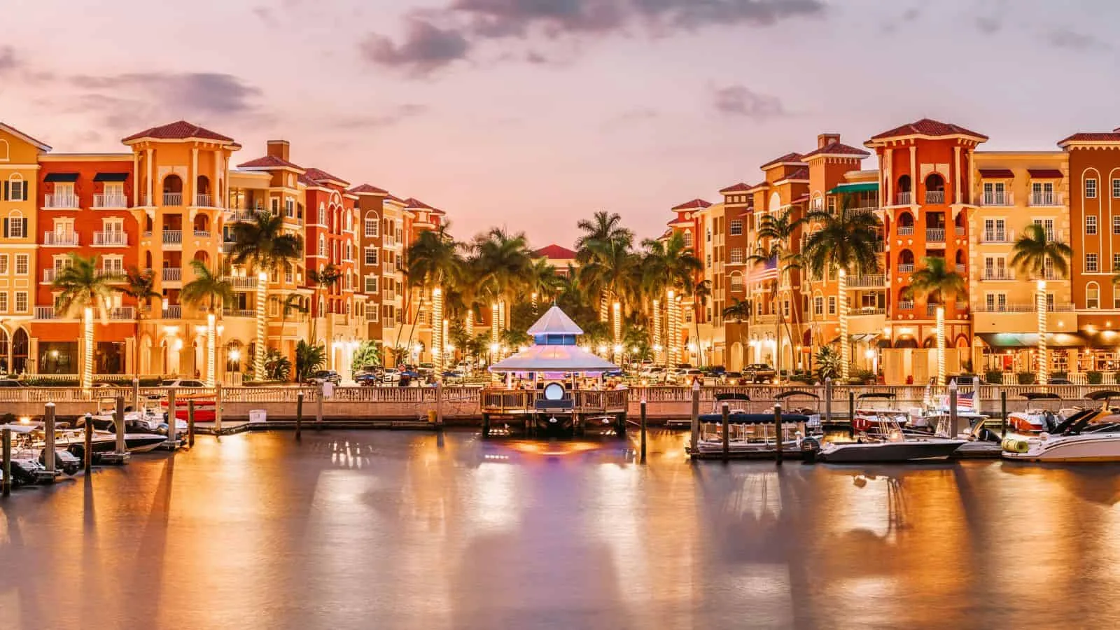 Waterfront residential plaza along Naples Bay at golden hour with a marina, docked boats, and a Mediterranean-style promenade