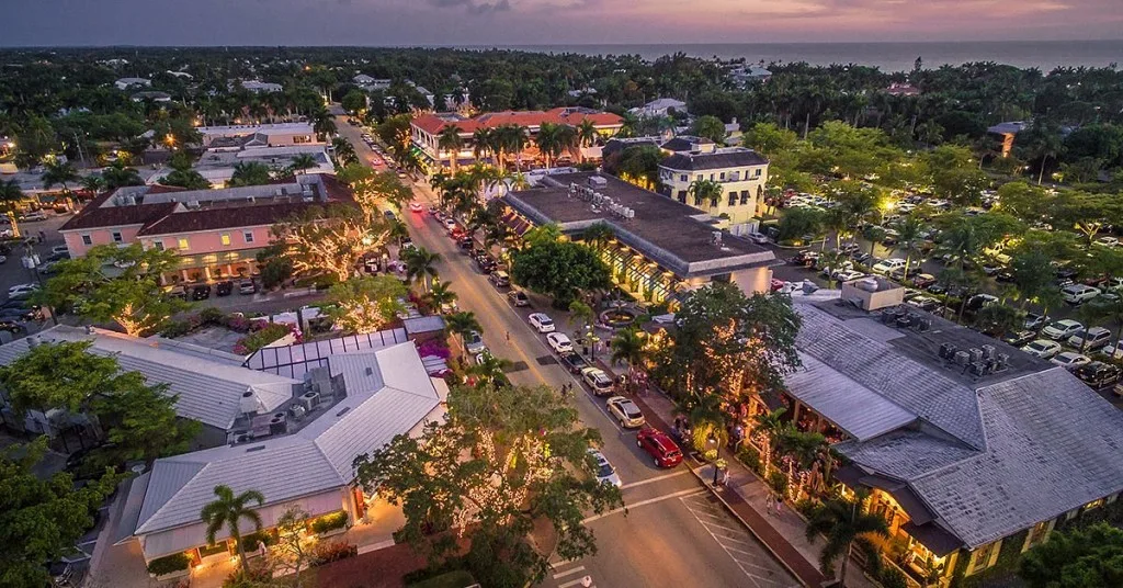 Twilight aerial of downtown Olde Naples with palm-lined streets and building lights glowing toward the Gulf