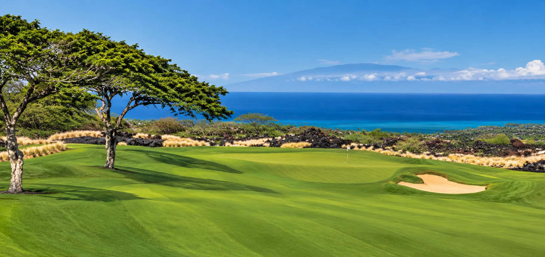 Manicured golf fairway with bunker and flag at Kūkiʻo Golf Club, surrounded by volcanic terrain with the ocean and a mountain peak in the distance.