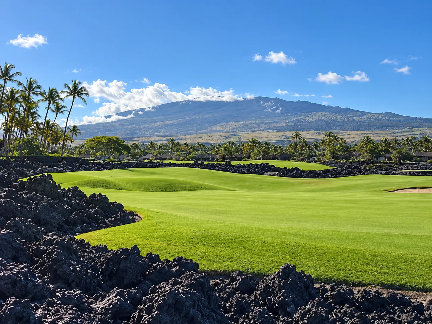 A lush green golf fairway bordered by dark lava rock and palms, with a large distant mountain under a blue sky