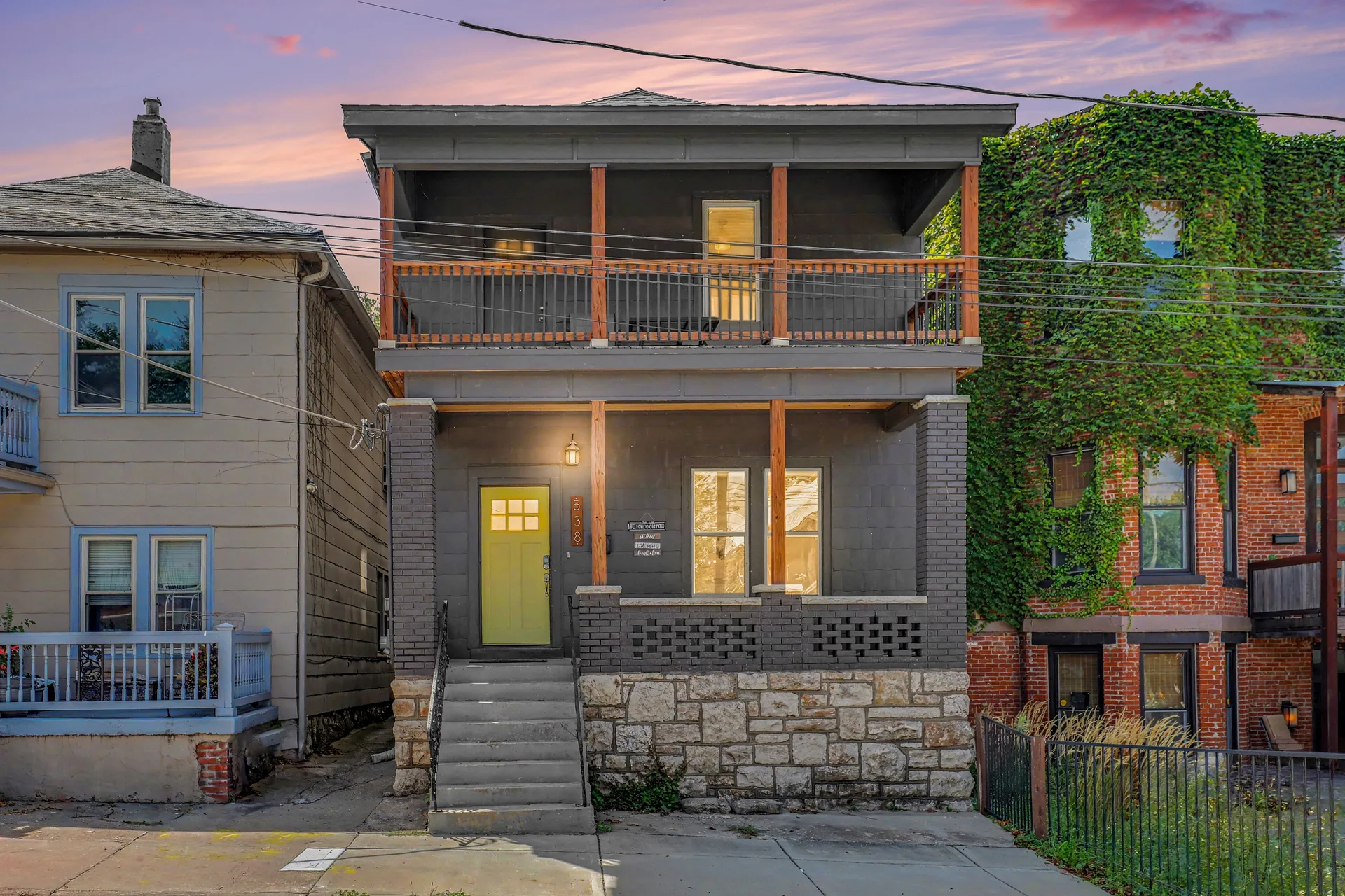 Front facade of 538 Harrison Street at sunset, a two-story urban townhome with dark gray painted brick, stone foundation, and wood-post porches with warm interior lights on