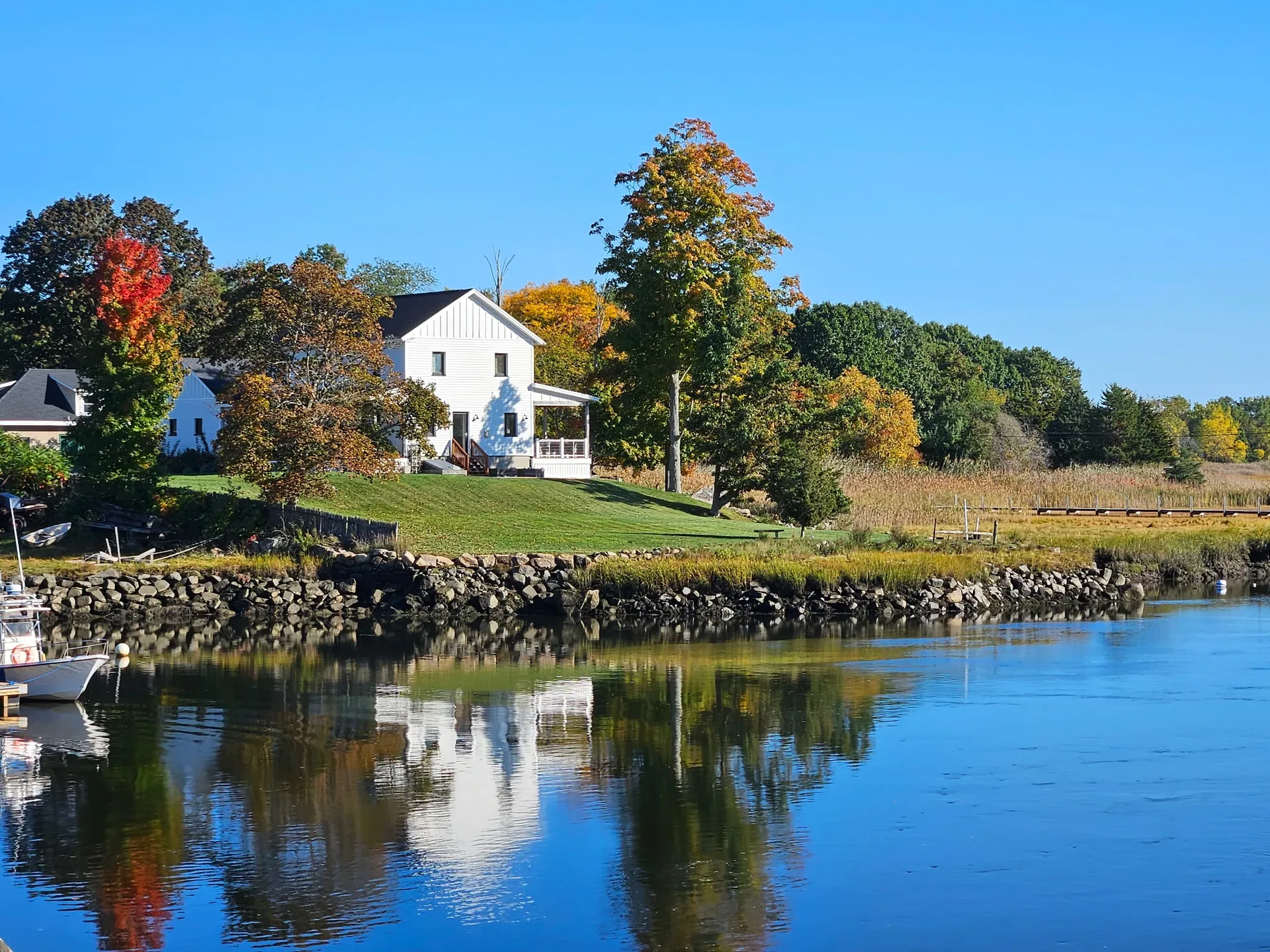 A white two-story house with a porch on a green lawn beside calm water, autumn trees in yellow, orange, and red, and a small boat on the left under a blue sky