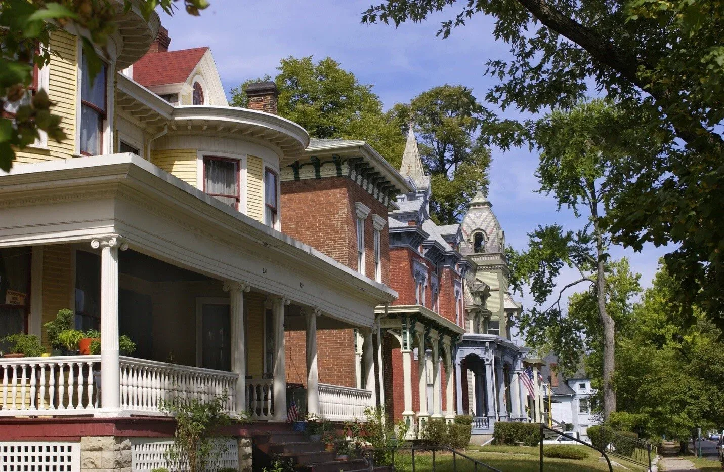 A tree-lined street of historic early-1900s homes on a bright autumn day, wood-sided houses with deep front porches and brick facades beneath tall mature trees turning orange and gold