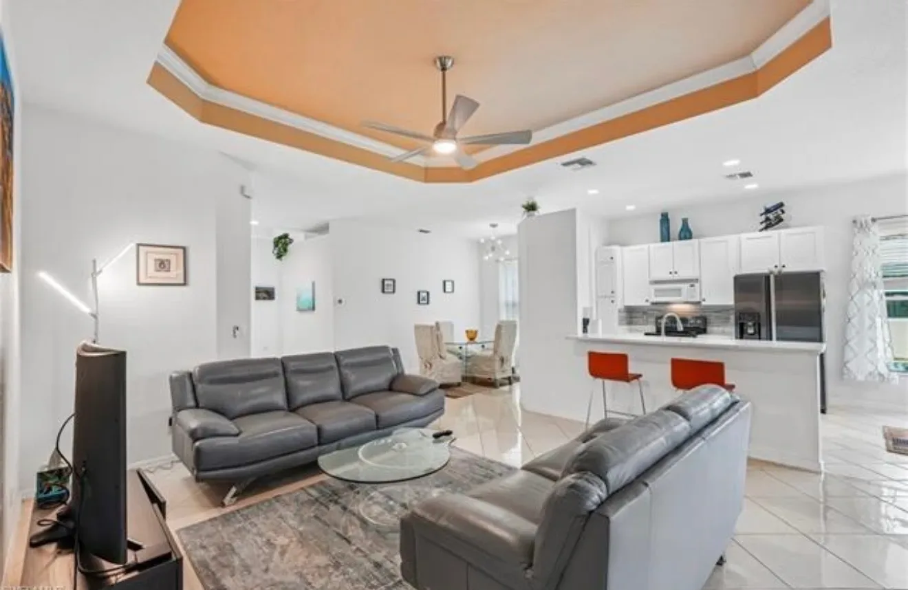 Open-concept living room and kitchen at 309 Harvard Ln, Naples, with grey leather sofas, a glass coffee table, white cabinets, and a breakfast bar with red stools