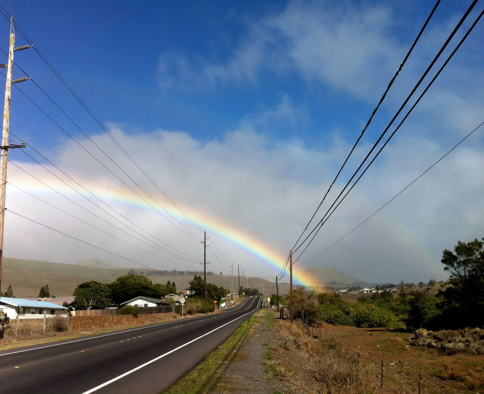 A two-lane road climbing from the coast toward green hills, with a rainbow stretching across the sky