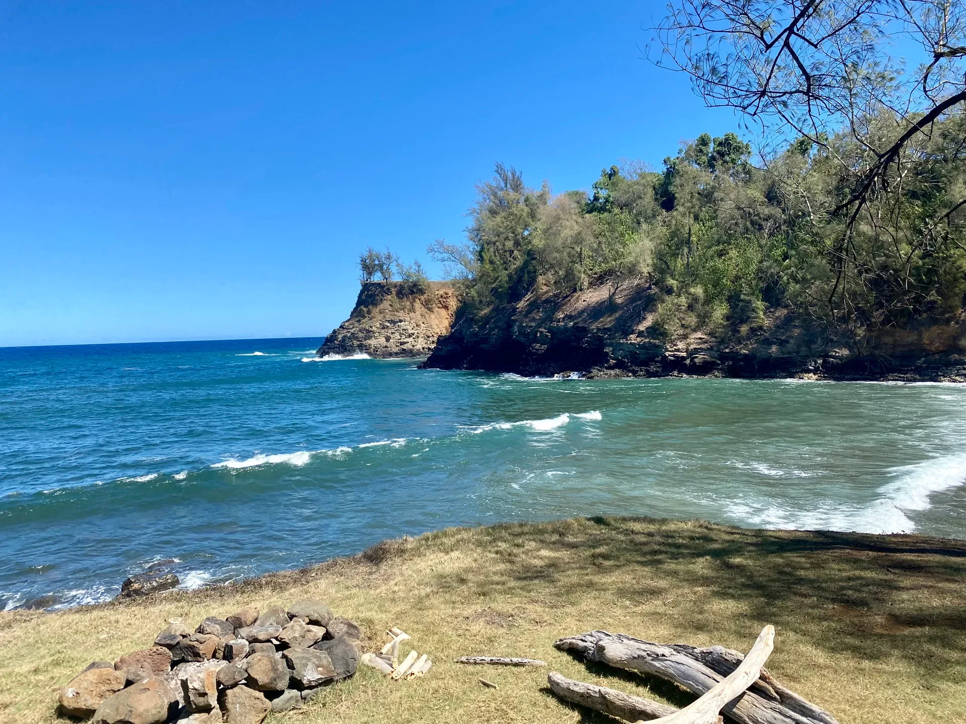 A grassy nook with a small stone fire pit on the rugged North Kohala coastline north of Kapaʻau, where a sheltered bay is framed by cliffs and green foliage