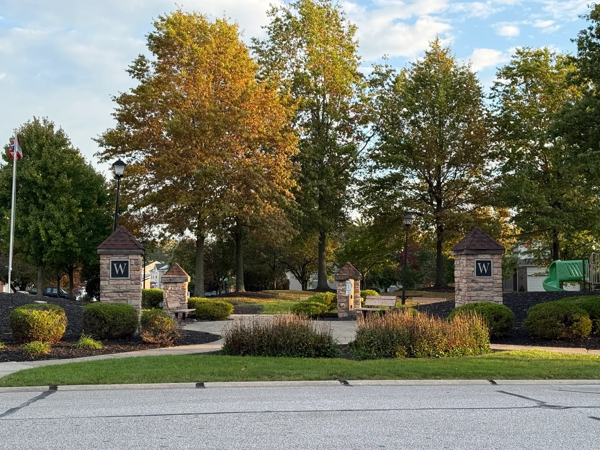 Neighborhood community entrance with stone pillars, landscaping, and mature trees under a clear blue sky