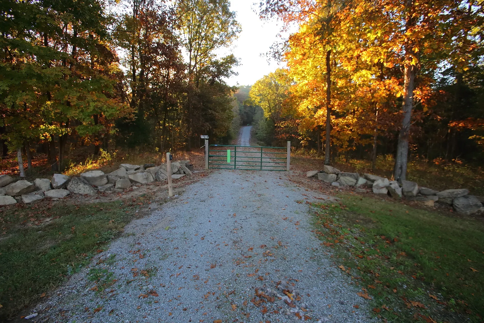 Gravel driveway entrance with a green metal gate framed by fieldstone, autumn woods