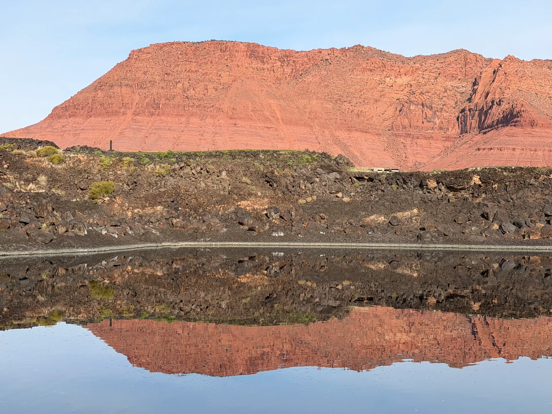 Red sandstone mesa reflecting in calm water at Black Desert Resort, with black lava rock in the foreground near Ivins, Utah