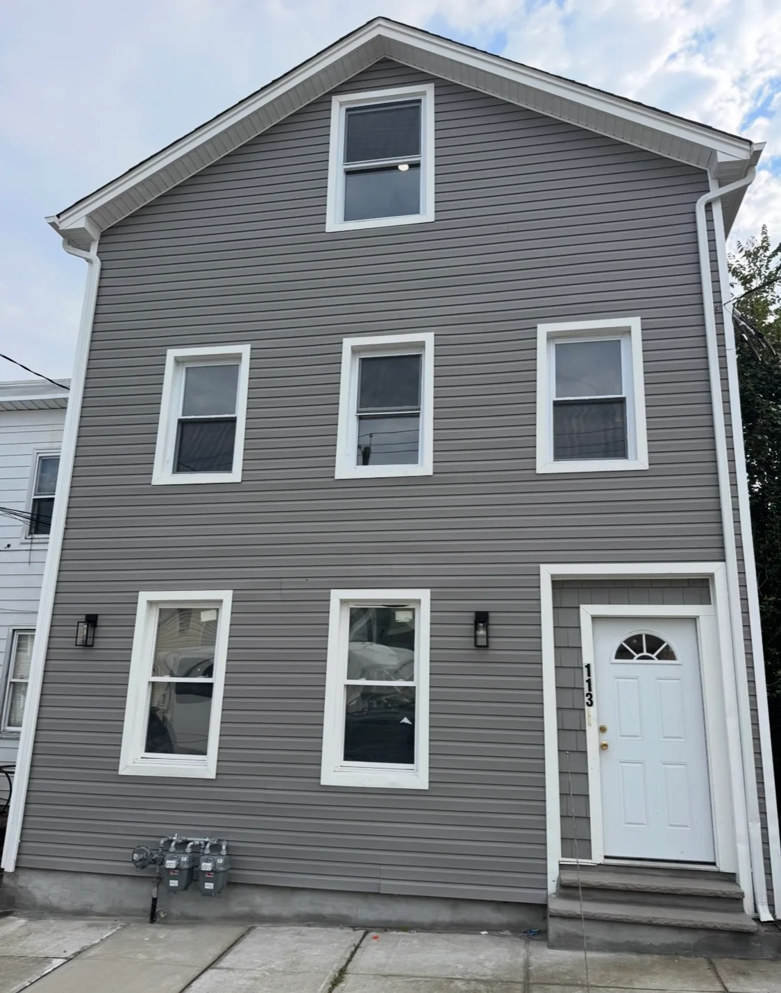 Three-story residential building front facade with gray vinyl siding, white trim, entry door, and concrete steps.