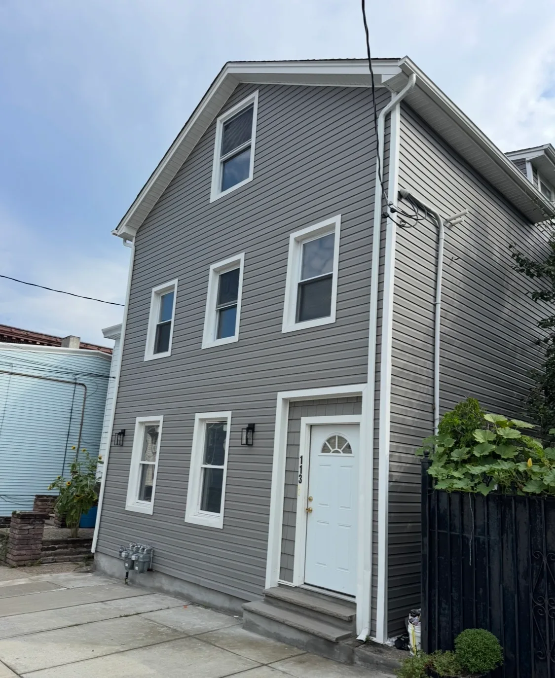 Front facade of a multi-story building with gray vinyl siding, white trim, utility meters, and a black fence.