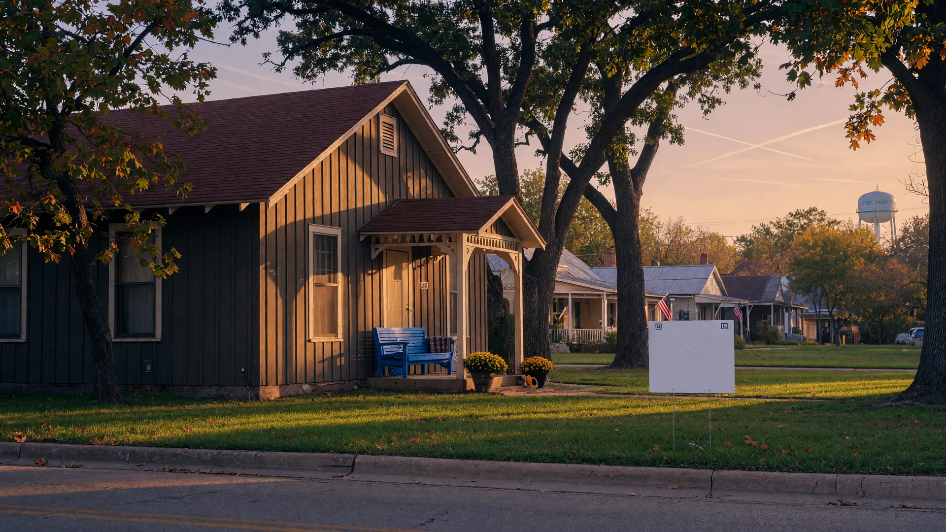 Small-town Texas home with dark siding, a covered porch, and a yard sign on a tree-lined Main Street at golden hour in early autumn