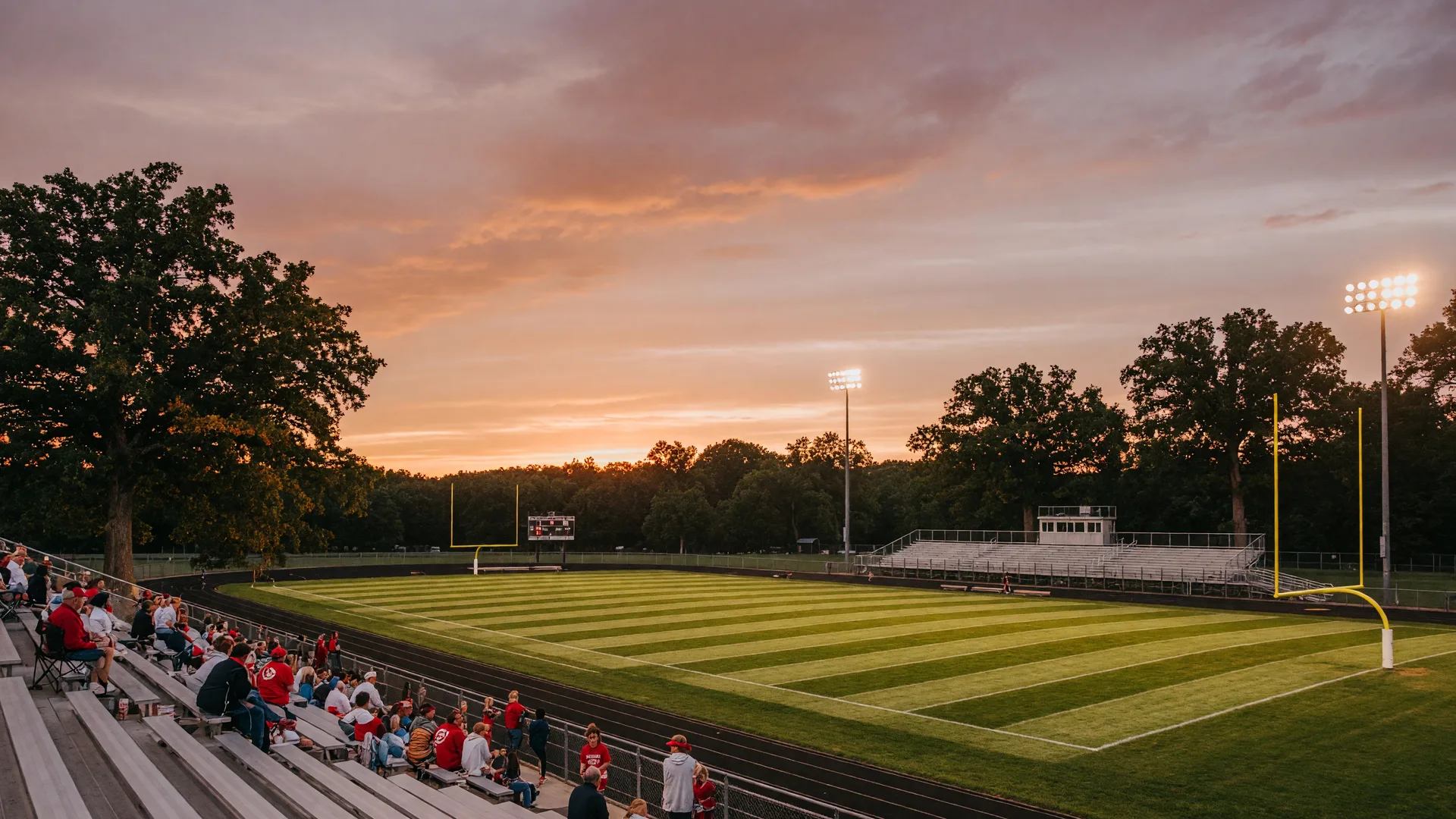 A rural Clinton County Ohio high school football stadium on a late summer evening with stadium lights glowing against a warm dusk sky, freshly striped field, and fans beginning to fill the bleachers