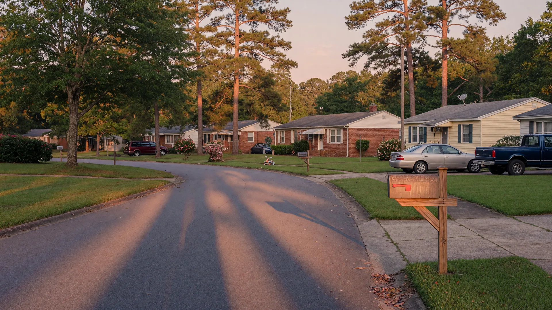 A quiet suburban street in the Columbia, SC Midlands in golden late-afternoon light, a wooden mailbox and well-kept homes under tall pines and oaks