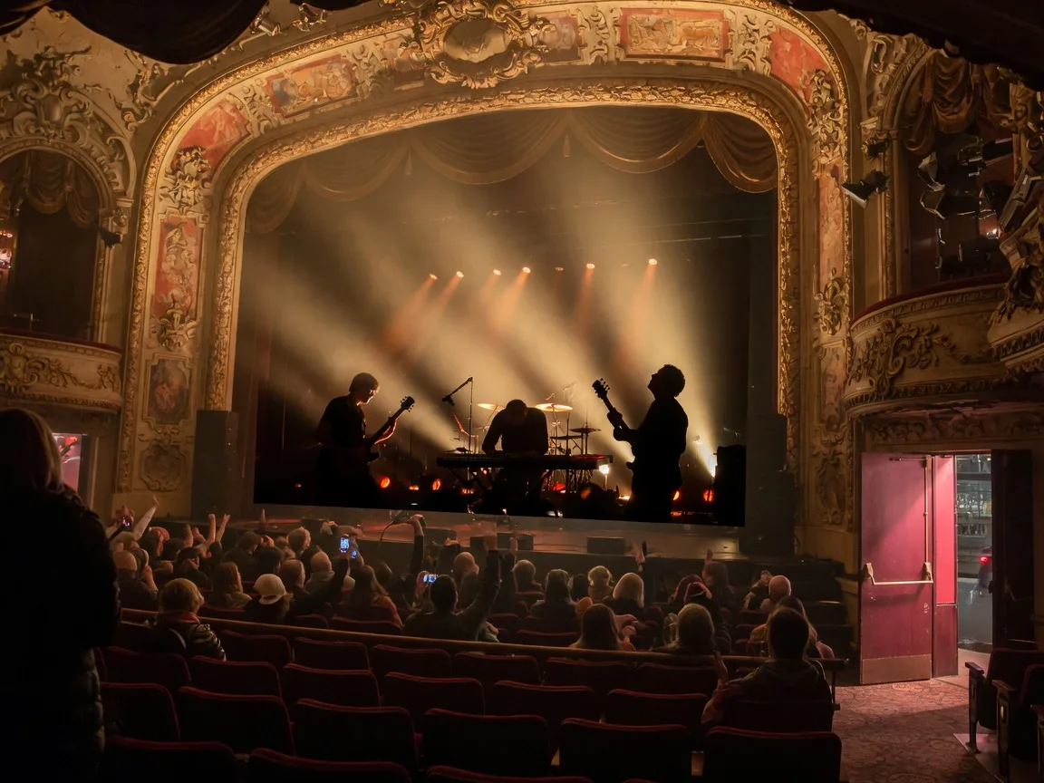 Warm amber stage light filling a historic 1920s theatre during a rock concert, a band as dark silhouettes on stage with guitars, haze and light beams rising to the ornate balconies, the front rows filled with a cheering audience