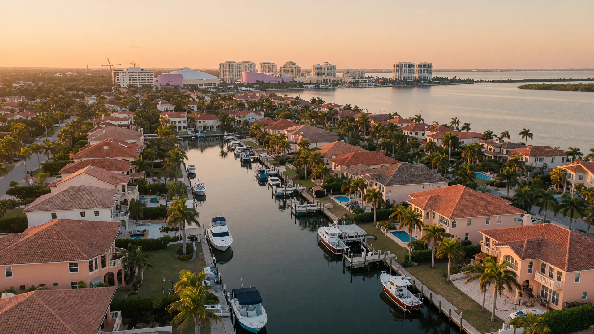 A low aerial view of an upscale Sarasota Bay neighborhood at golden hour, Mediterranean-style homes with terracotta roofs and private docks lining calm residential canals with moored boats, royal palms in warm amber light, downtown skyline soft in the distance
