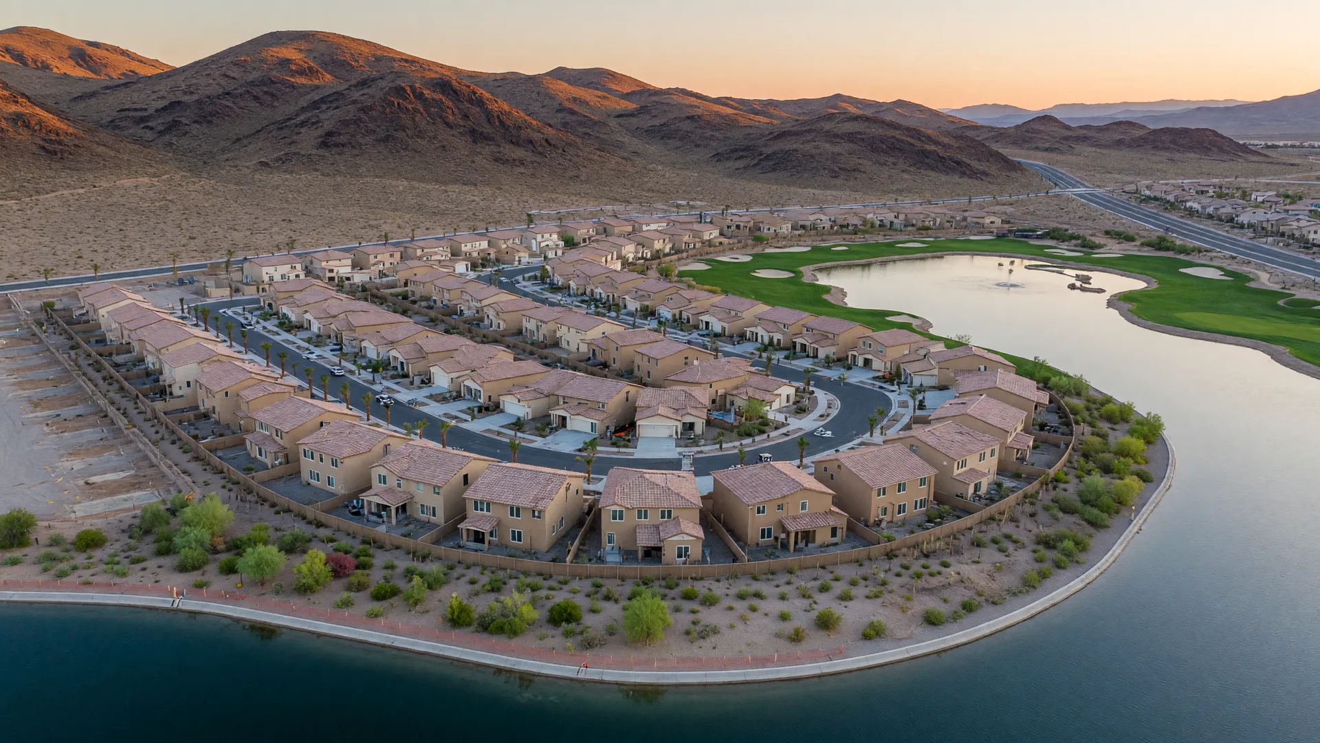 Aerial view of a new master-planned community of tan stucco homes and palm-lined streets near a desert lake with hills in the distance
