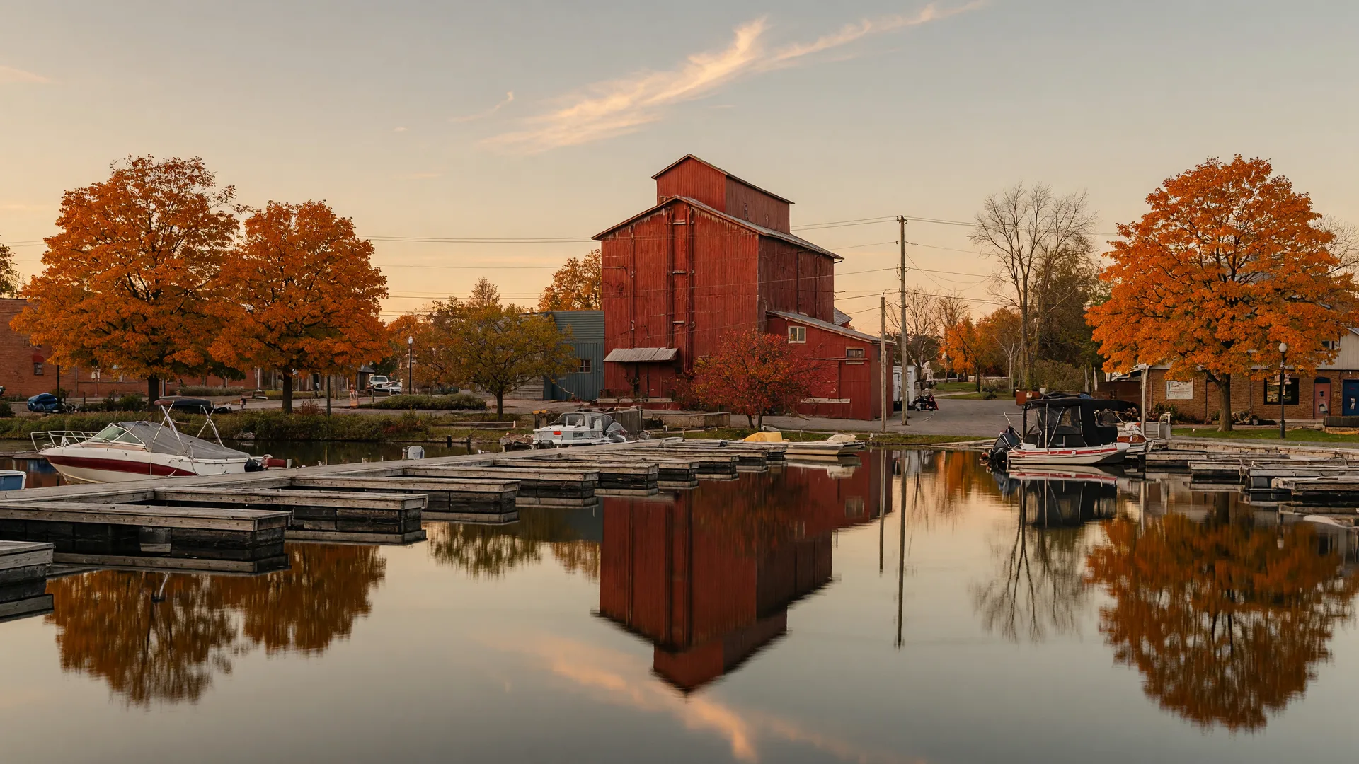 The Port Perry waterfront on Lake Scugog in late autumn, seen from the water with the heritage red brick grain mill at the foot of Queen Street, wooden marina docks with moored boats, and sugar maples in orange and gold along the shoreline