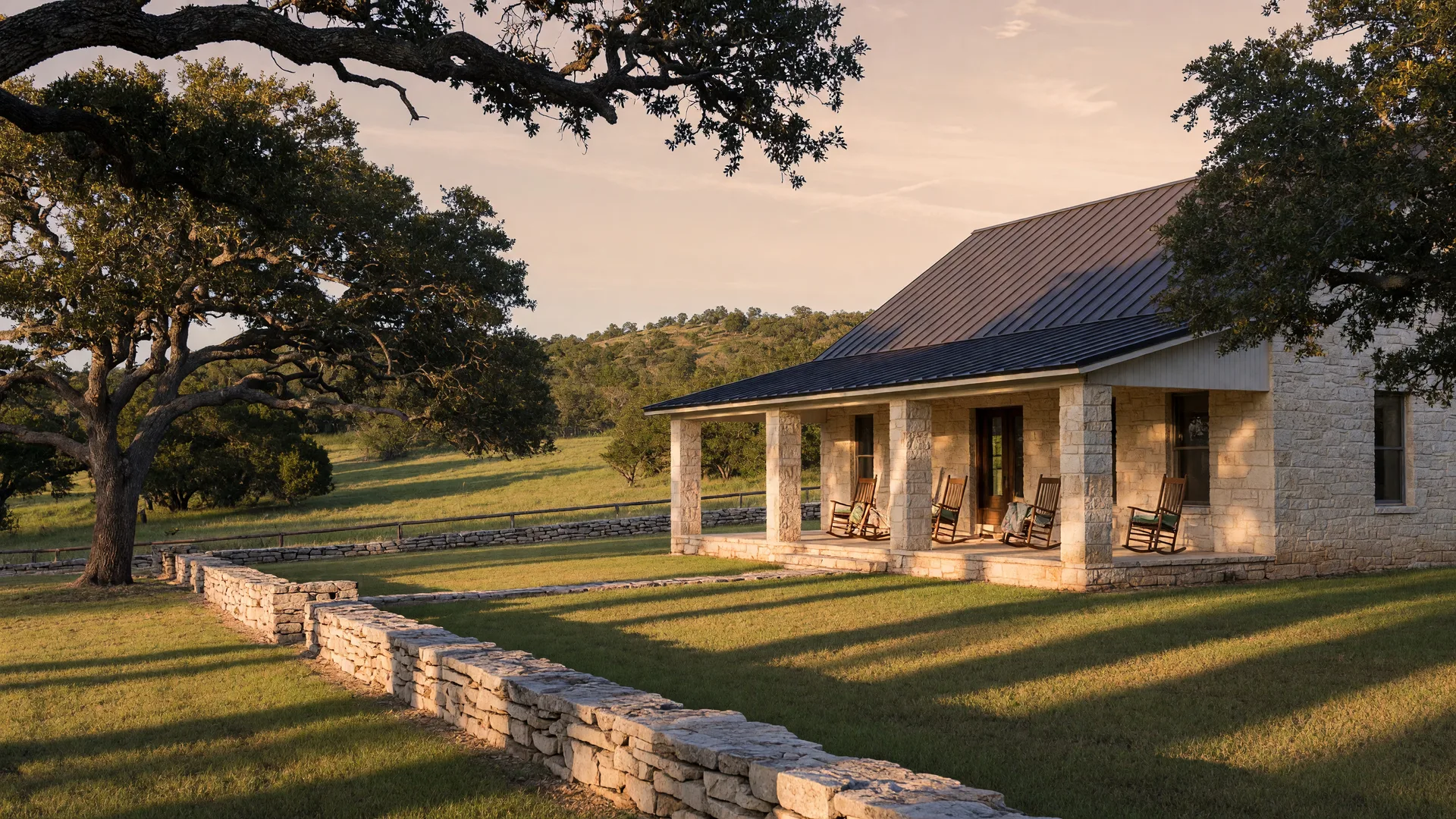 A limestone Hill Country home in Boerne with a metal roof, live oaks, and a stacked-stone fence in late afternoon light