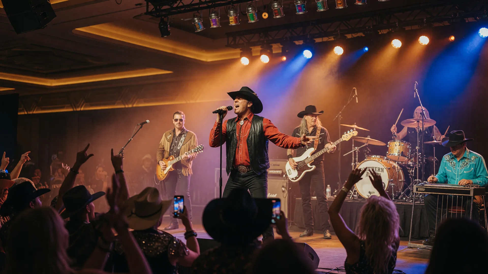 A country and southern rock band performing live on stage with amber and blue stage lighting and crowd silhouettes