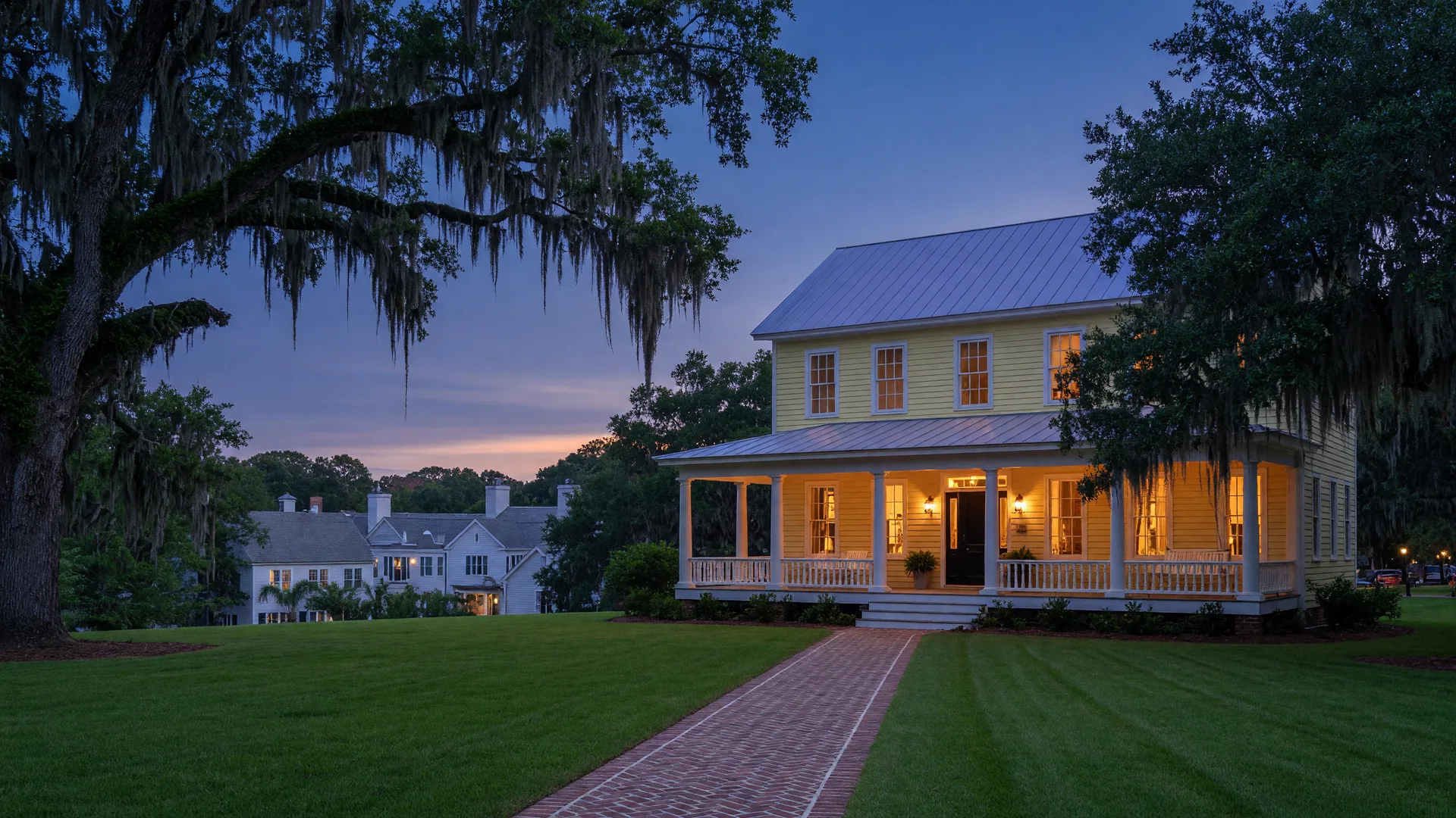 A Charleston Lowcountry home at dusk with warm lights glowing through the front windows, moss-draped live oaks framing the yard