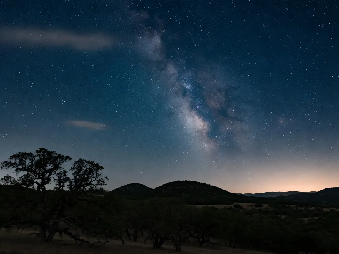 A star-filled Texas Hill Country night sky with the Milky Way arching over silhouetted live oaks and rolling hills
