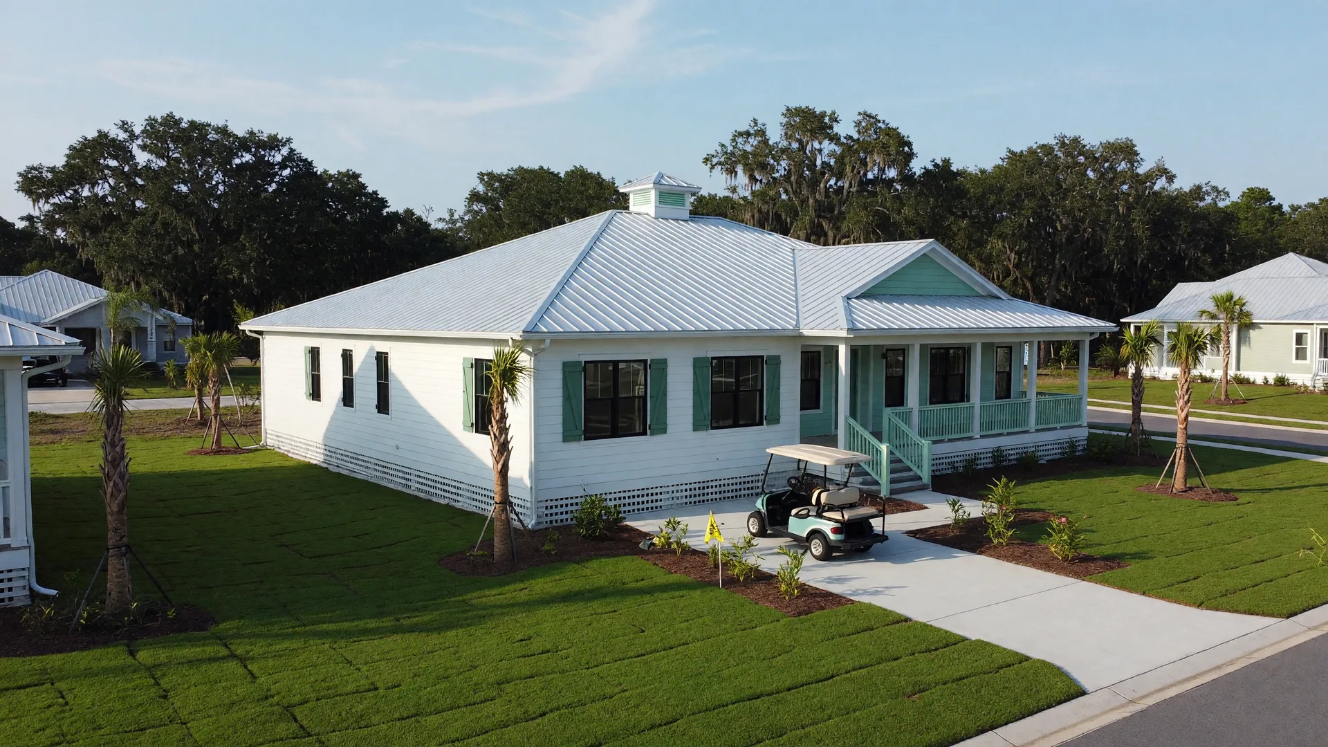 Brand-new single-story coastal home in a Gulf Coast subdivision near Orange Beach with a metal roof, impact windows, young palms, and a golf cart in the driveway