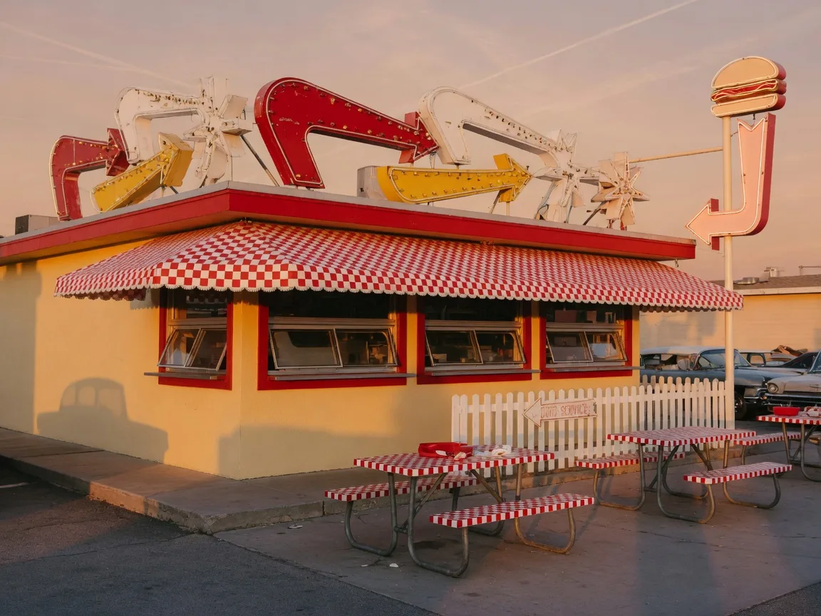 A retro drive-in burger stand with red and white awnings and picnic tables out front at golden hour, evoking The Big M Drive In in Pickering
