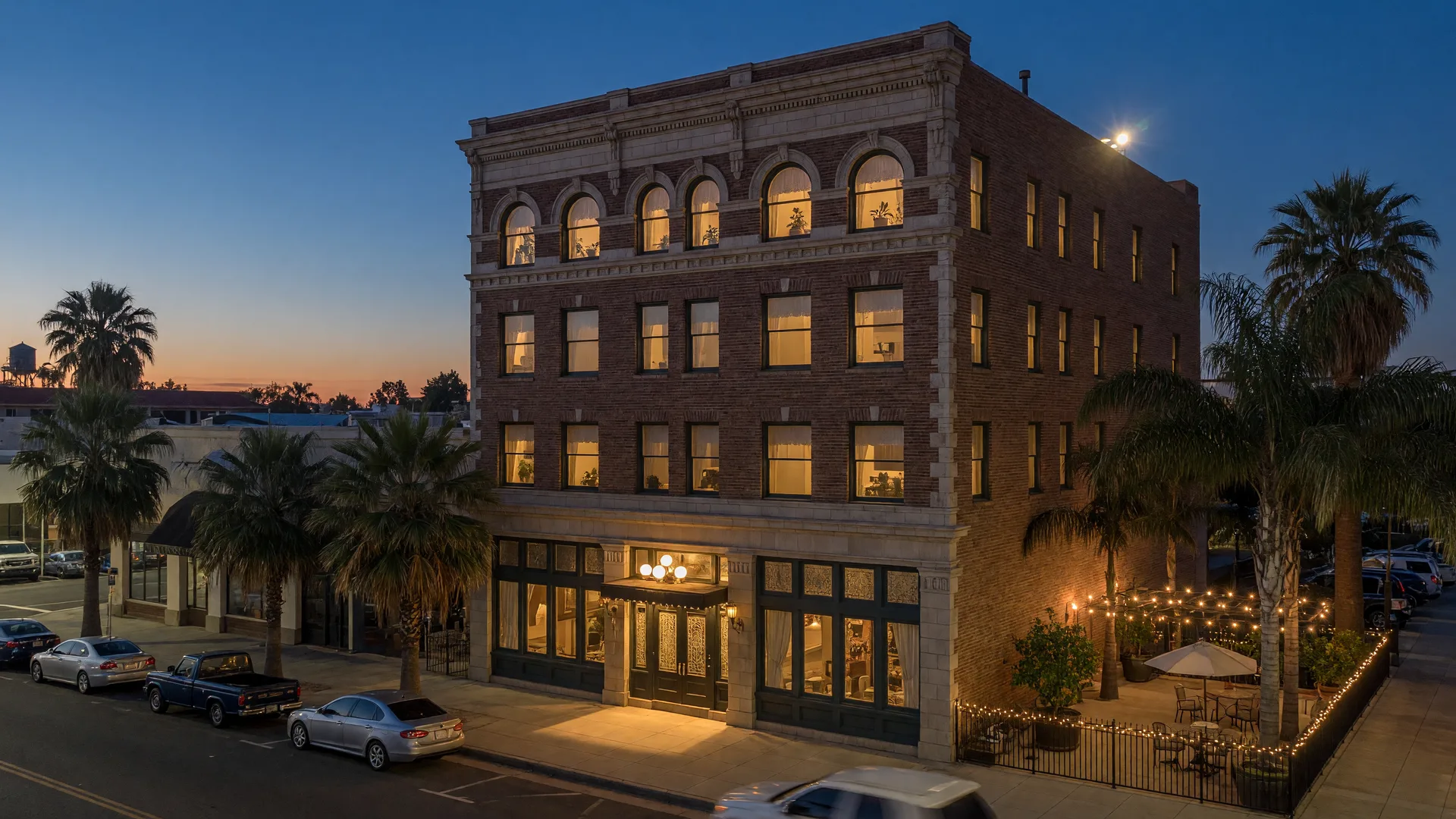 The restored four-story El Capitan Hotel on West Main Street in downtown Merced at dusk, with warmly glowing arched windows and palm trees along the street