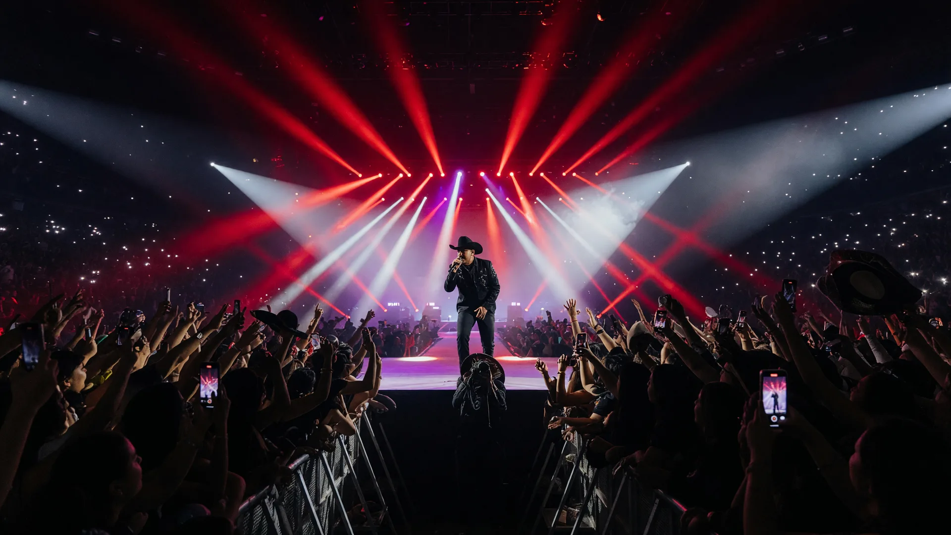 A regional Mexican corridos concert with a singer in a hat at center stage under red and white light beams and a huge silhouetted crowd