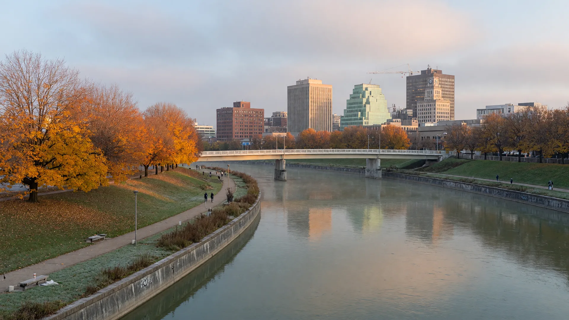 The Great Miami River flowing through downtown Dayton on a crisp late-October morning, calm water between low floodwall banks, a pedestrian footbridge spanning the river, and the brick and mid-rise skyline rising beyond golden autumn trees
