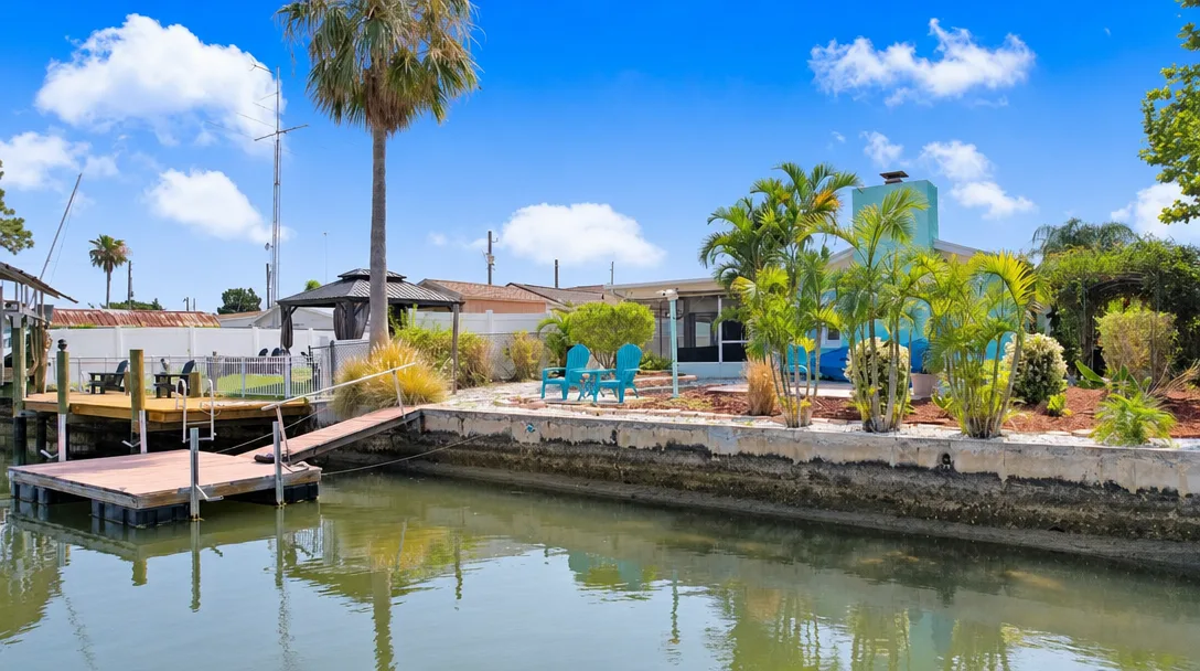 A Hudson, Florida canal-front ranch home on a clear morning, with a private dock, a small boat tied alongside, a seawall along the waterfront yard, and palm trees framing the scene