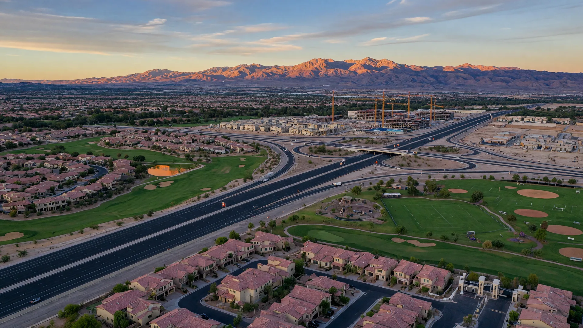 Aerial golden-hour view of Southwest Las Vegas master-planned communities, a guard-gated golf course, the 215 Beltway with new on-ramps, and tower cranes over new construction in mid-October
