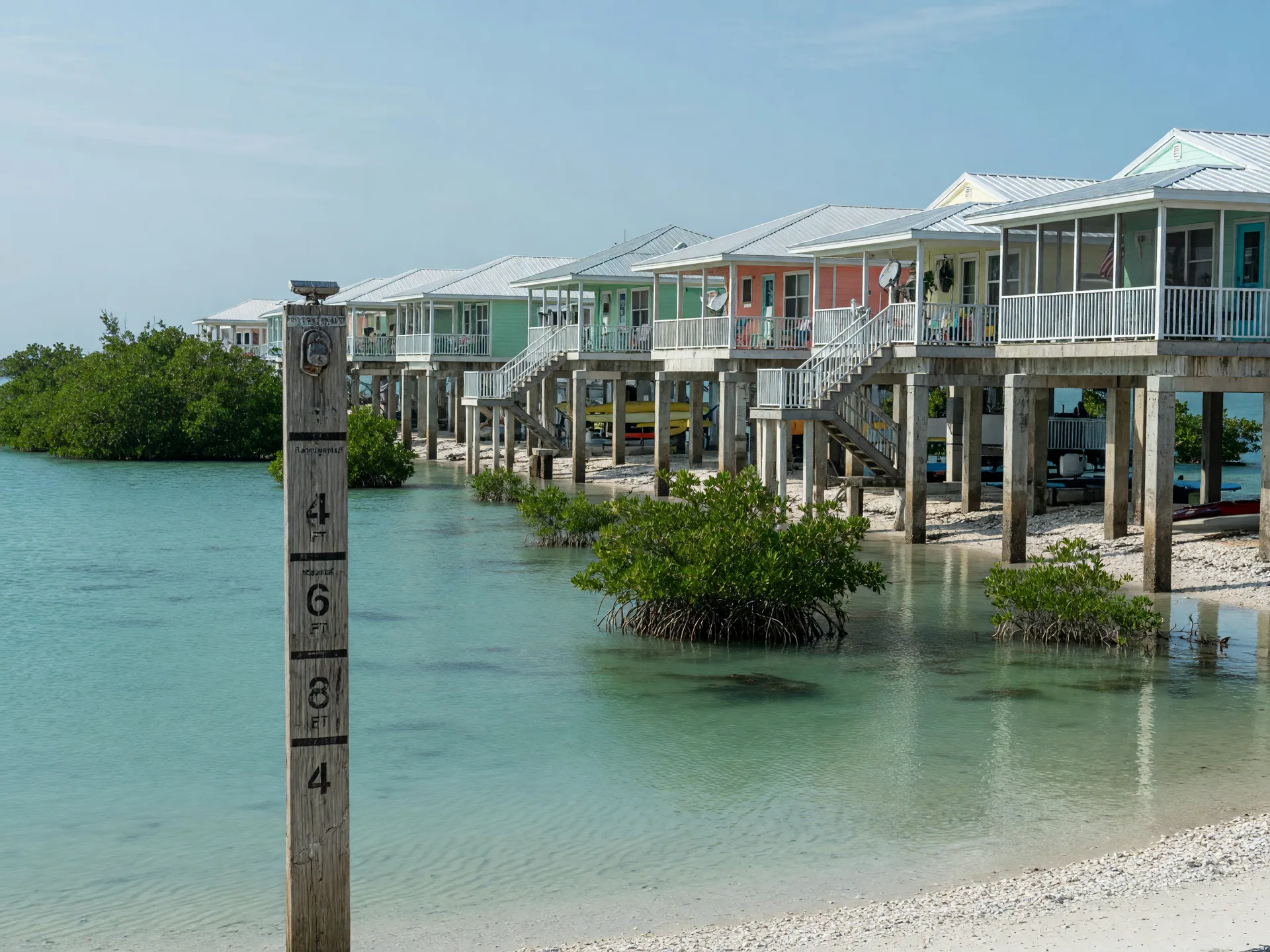 Florida Keys homes elevated on pilings along a calm coastal shoreline with a wooden flood elevation marker in the foreground