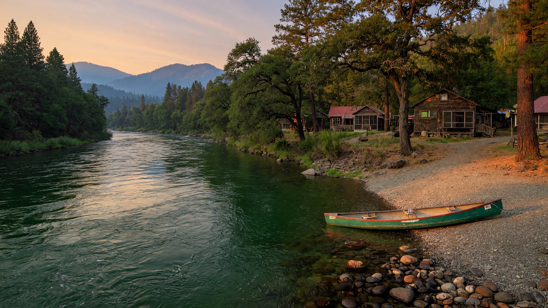 The upper Rogue River sliding past Shady Cove in warm evening light, a canoe at the gravel launch and cabins among the pines and oaks