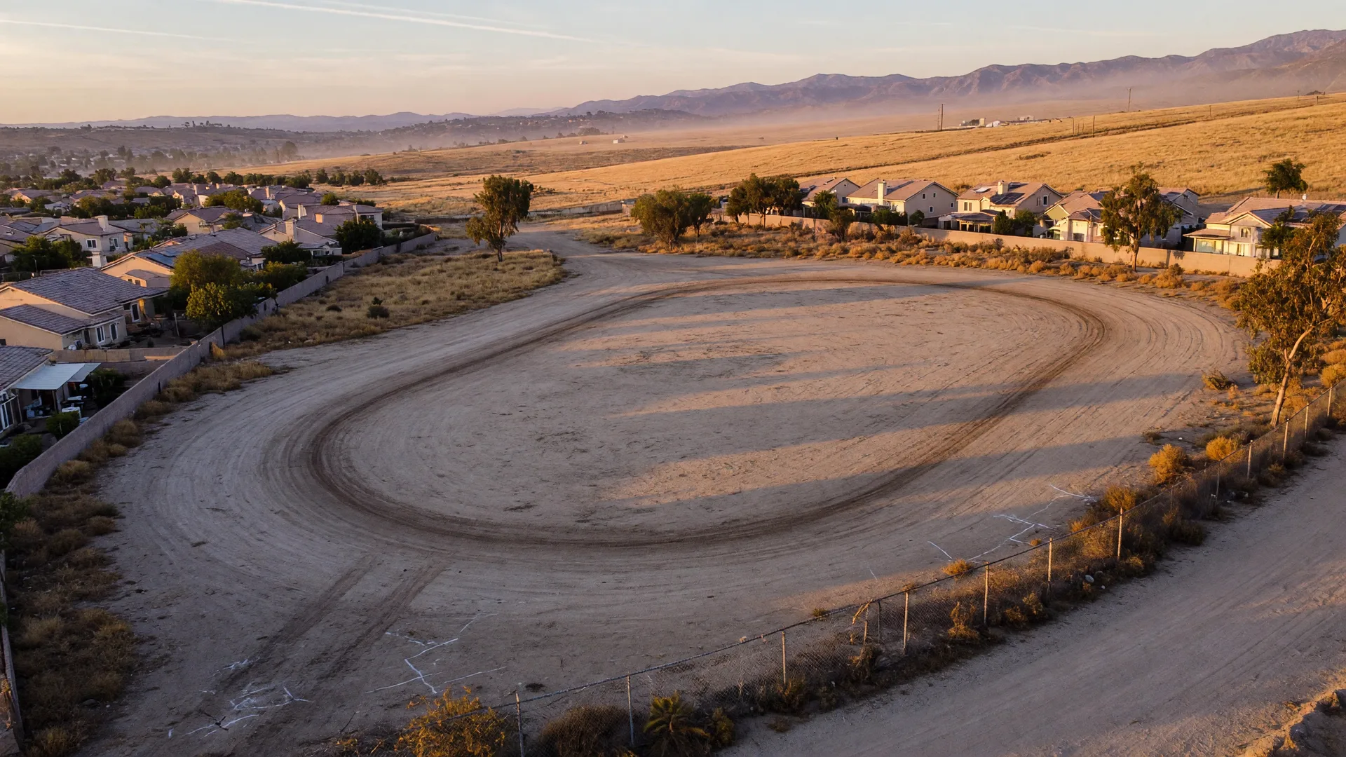 The cleared former Saugus Speedway lot at golden hour with the old oval track outline in the dirt and mountains beyond