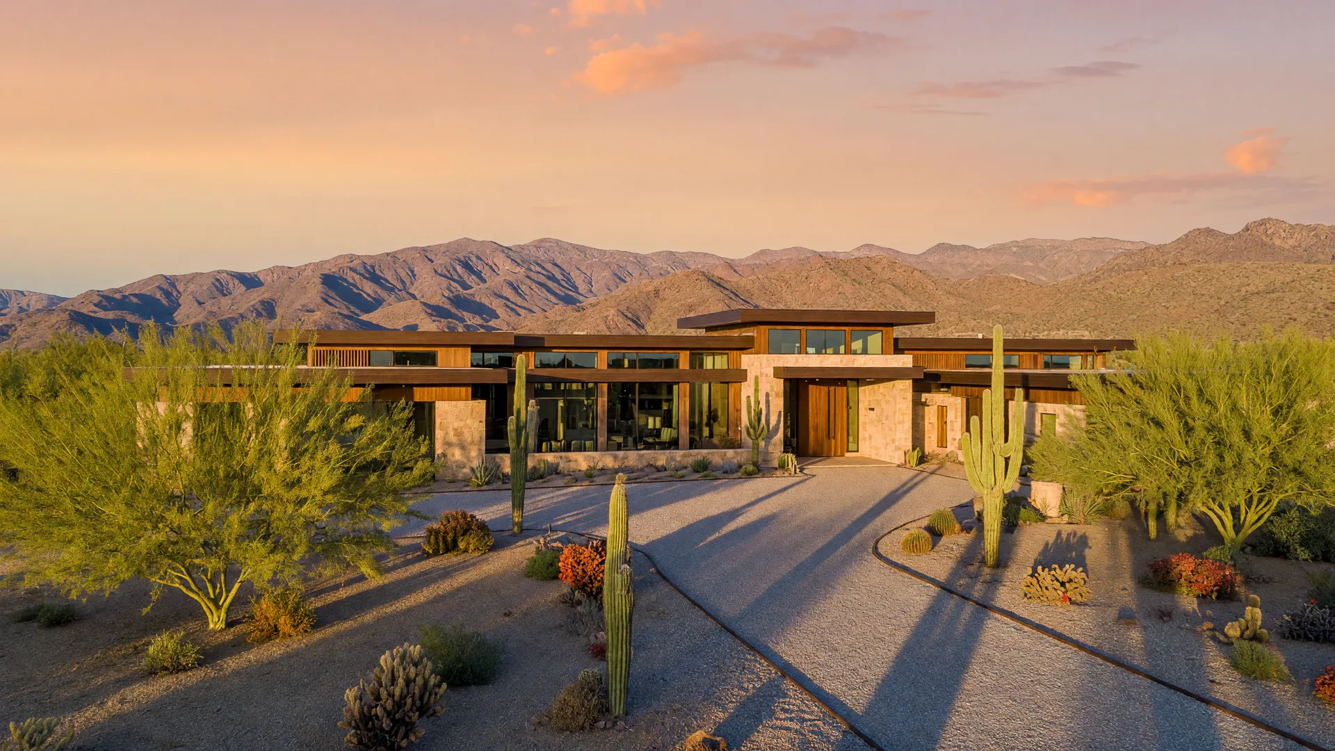 A modern desert estate on a large Queen Creek lot at golden hour, floor-to-ceiling glass glowing under palo verde trees and saguaros with the San Tan Mountains behind