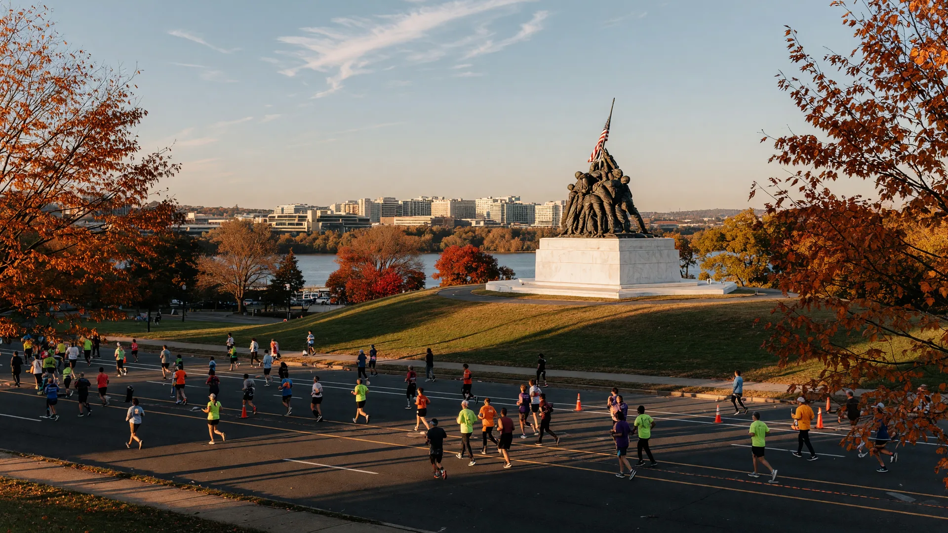 Wide view of the Marine Corps Marathon finish area in the Rosslyn neighborhood of Arlington, Virginia on a late-October morning, distant runners crossing Route 110 near the Pentagon in golden low sunlight, the Marine Corps War Memorial on its grassy hill, and autumn trees in red and orange