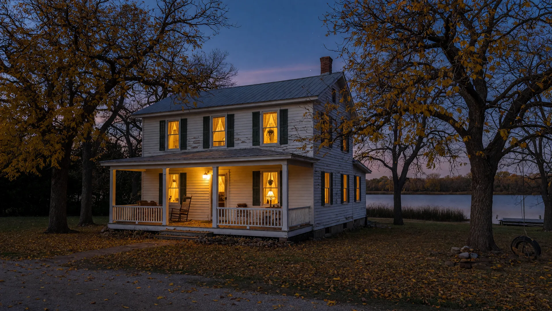 A historic white farmhouse with a covered front porch at dusk in late autumn, warm lamplight glowing from the windows, golden pecan trees around it and calm lake water in the distance