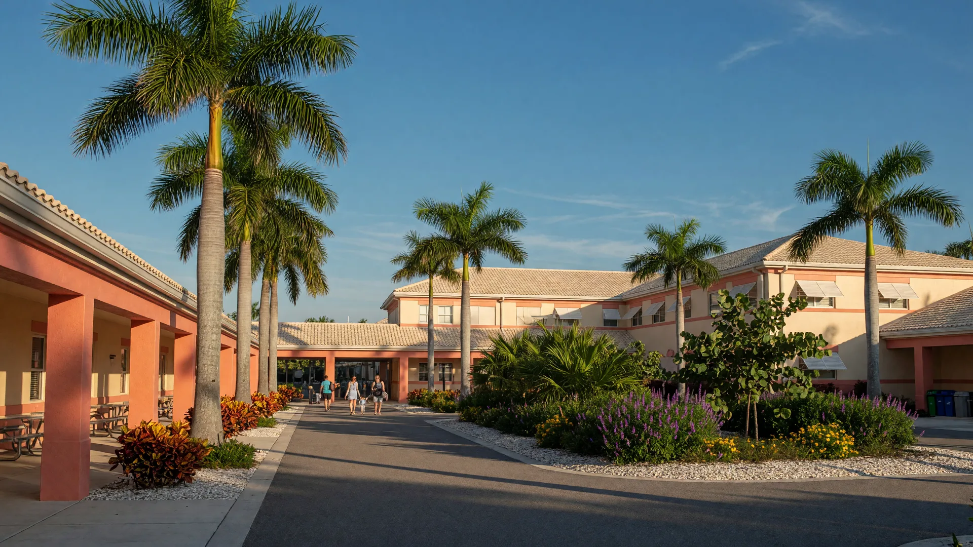 Modern public school campus in Naples, Florida with low-rise stucco buildings, royal palms along the entry drive, and parents walking children to class on a sunny morning
