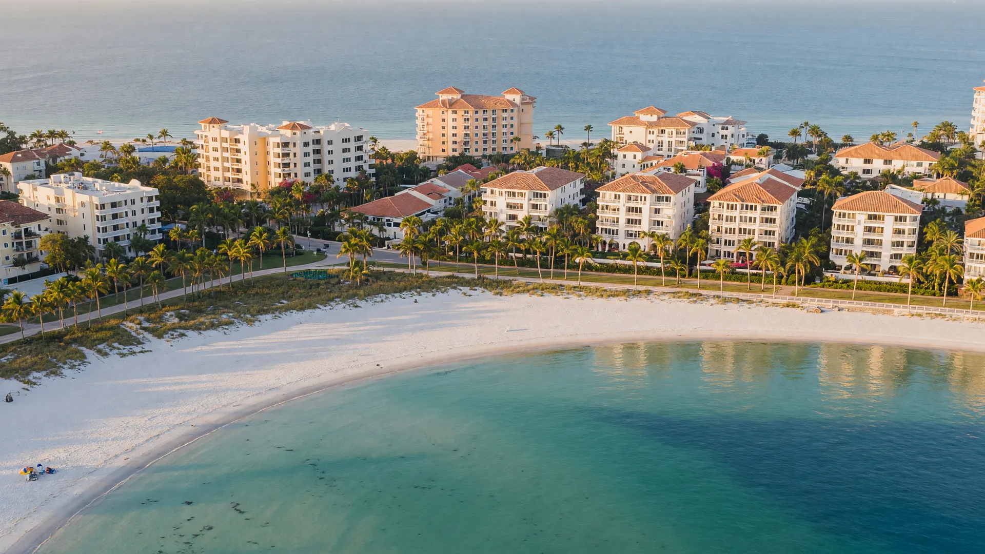 Aerial view of an elegant Gulf Coast Florida retirement city at golden hour, with turquoise bay water, Mediterranean-style stucco condominiums, terracotta roofs, palm-lined boulevards, and a curving white sand beach