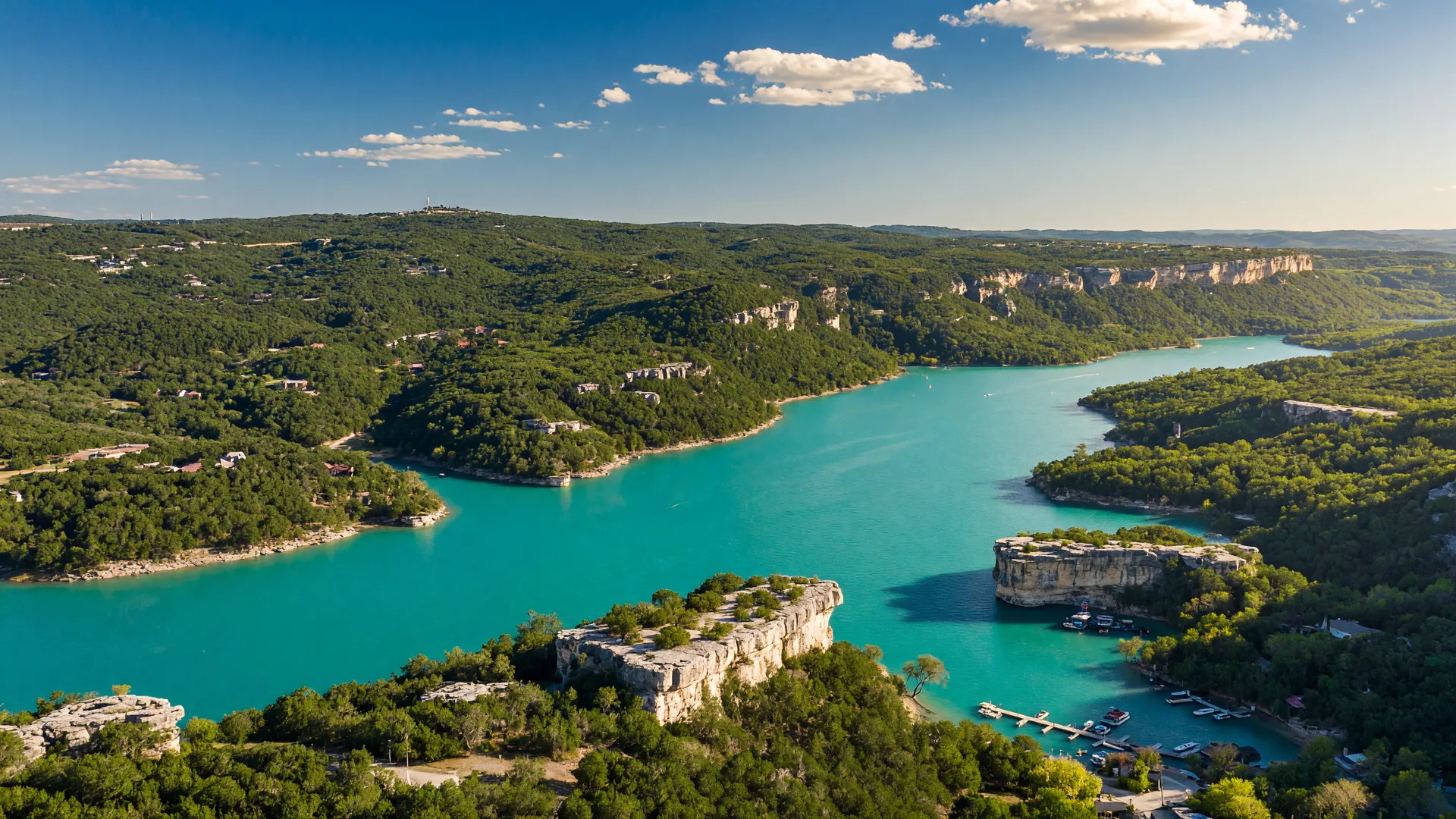 Aerial panorama of Canyon Lake in the Texas Hill Country — brilliant turquoise water surrounded by rolling green hills and limestone bluffs under a sunny blue sky