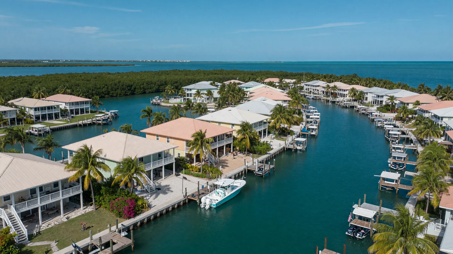 Aerial view of stilt-style waterfront homes lining a deep turquoise canal in the Florida Keys, with private docks, palm trees, and open water beyond