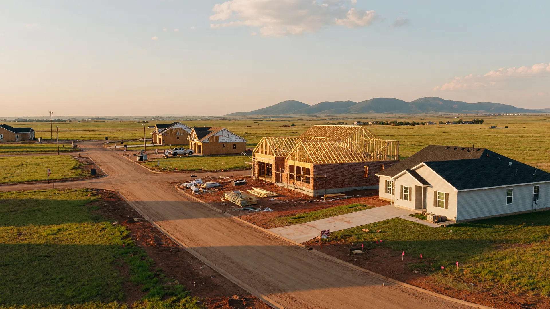A new single-family home under construction on a large grassy homesite in an Elgin, Oklahoma addition at golden hour, with timber framing and fresh brick beside a finished home and the Wichita Mountains on the horizon